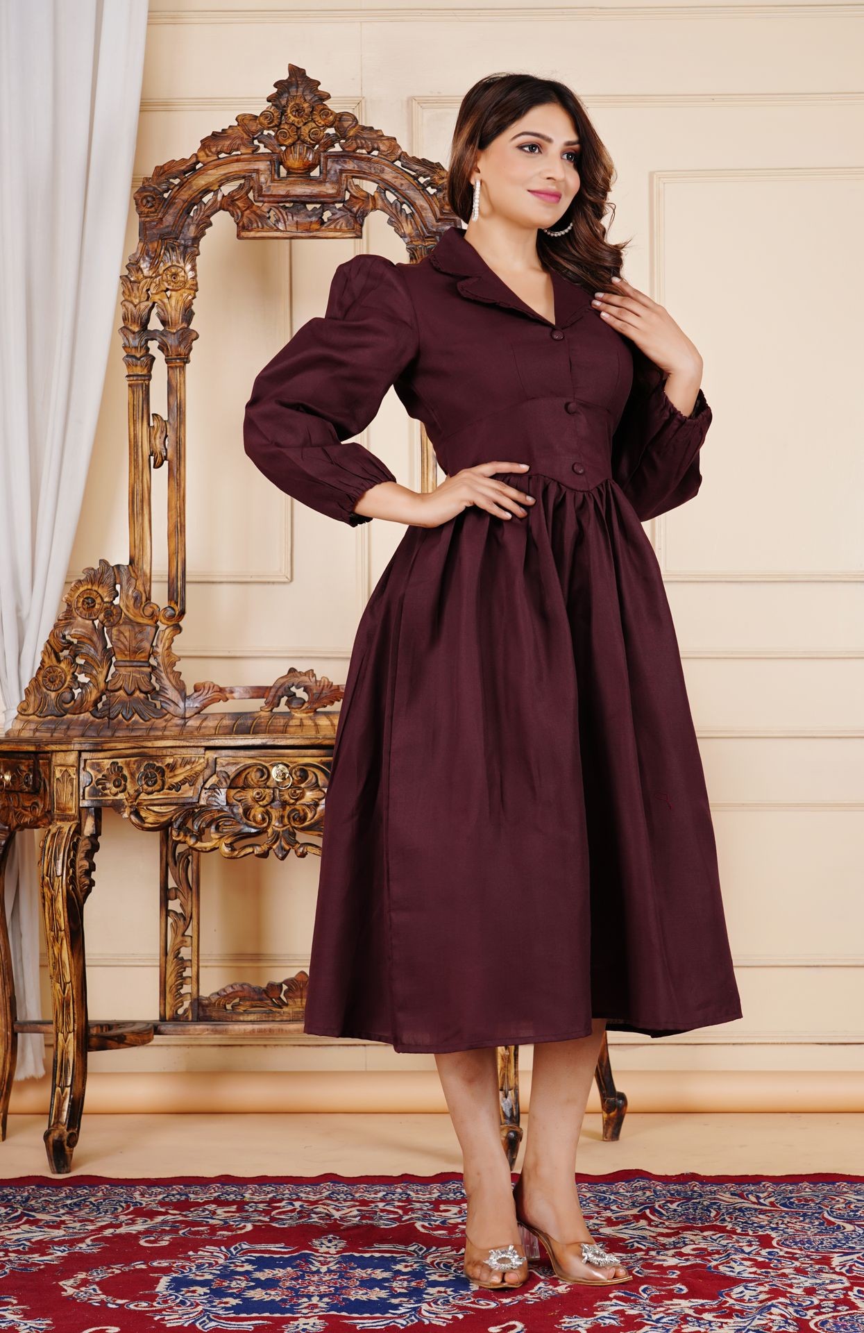 Woman in maroon dress poses by ornate wooden chair on patterned rug.