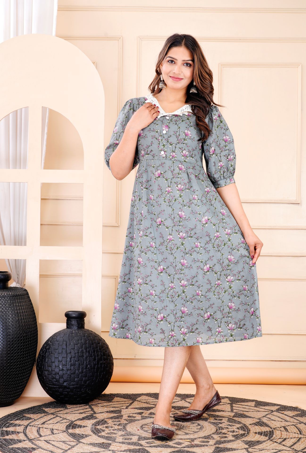 Woman in a floral printed dress posing indoors with decorative vases and a patterned rug.