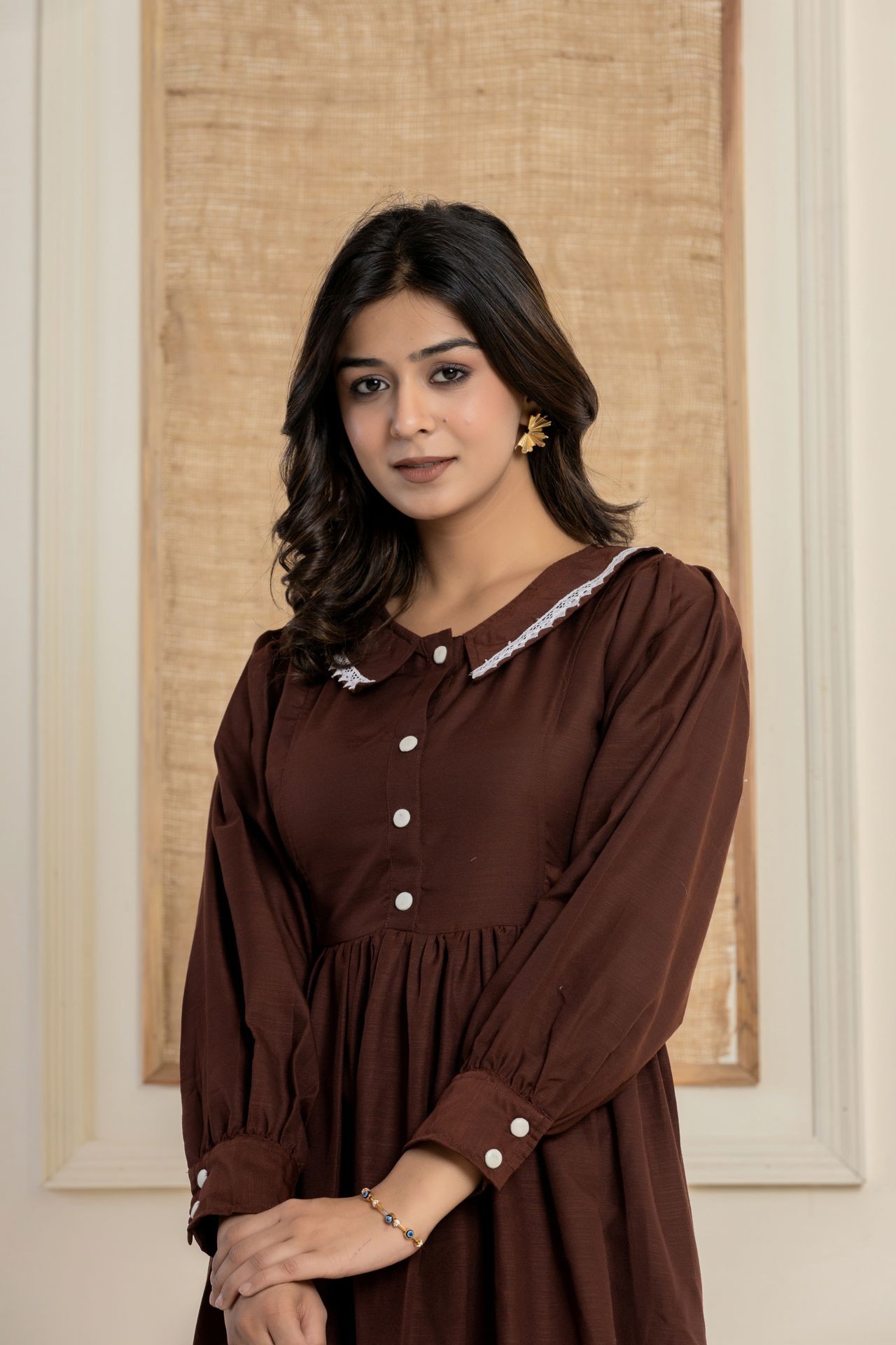 Woman in brown dress with white buttons, standing indoors with a neutral expression.