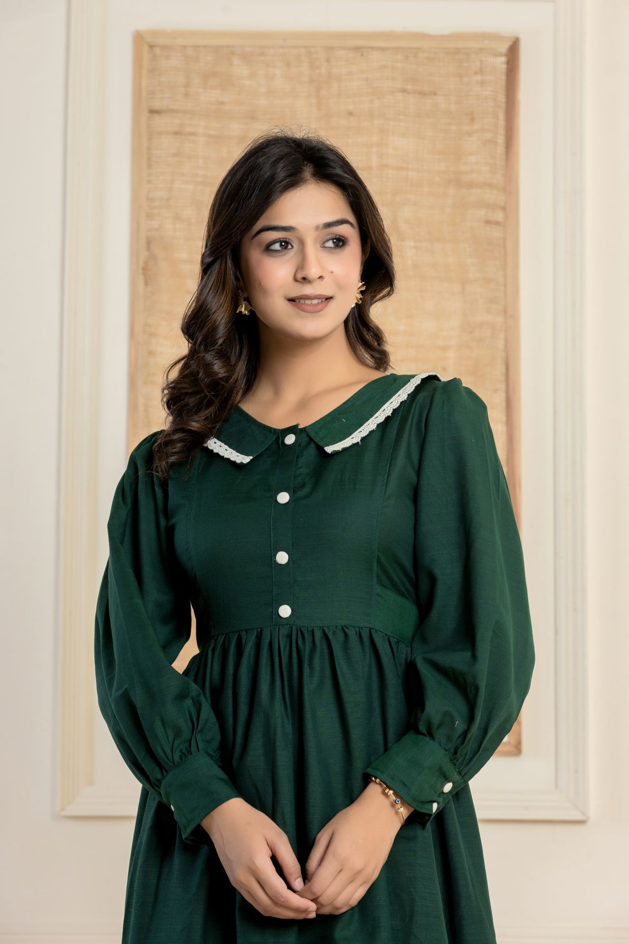 Woman in a green dress with white buttons and lace collar, standing indoors against a textured background.