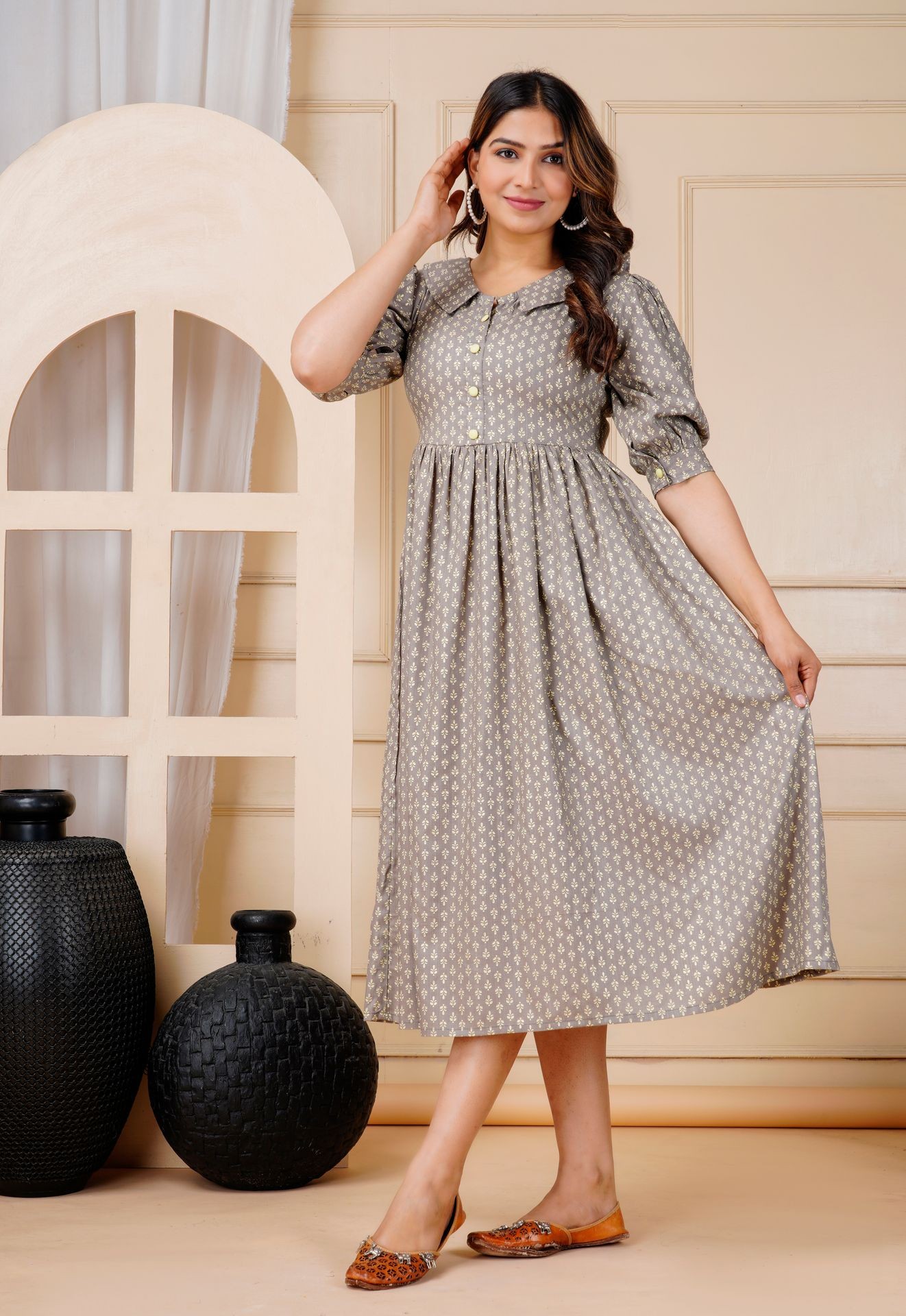 Person wearing a long, patterned dress stands near decorative vases and a window-like backdrop.