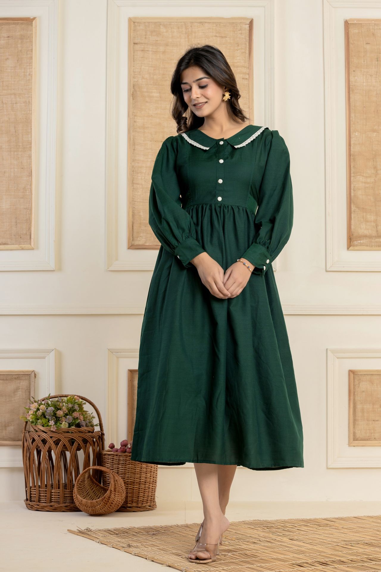 Woman wearing a long green dress with buttons, standing indoors near wicker baskets filled with flowers.