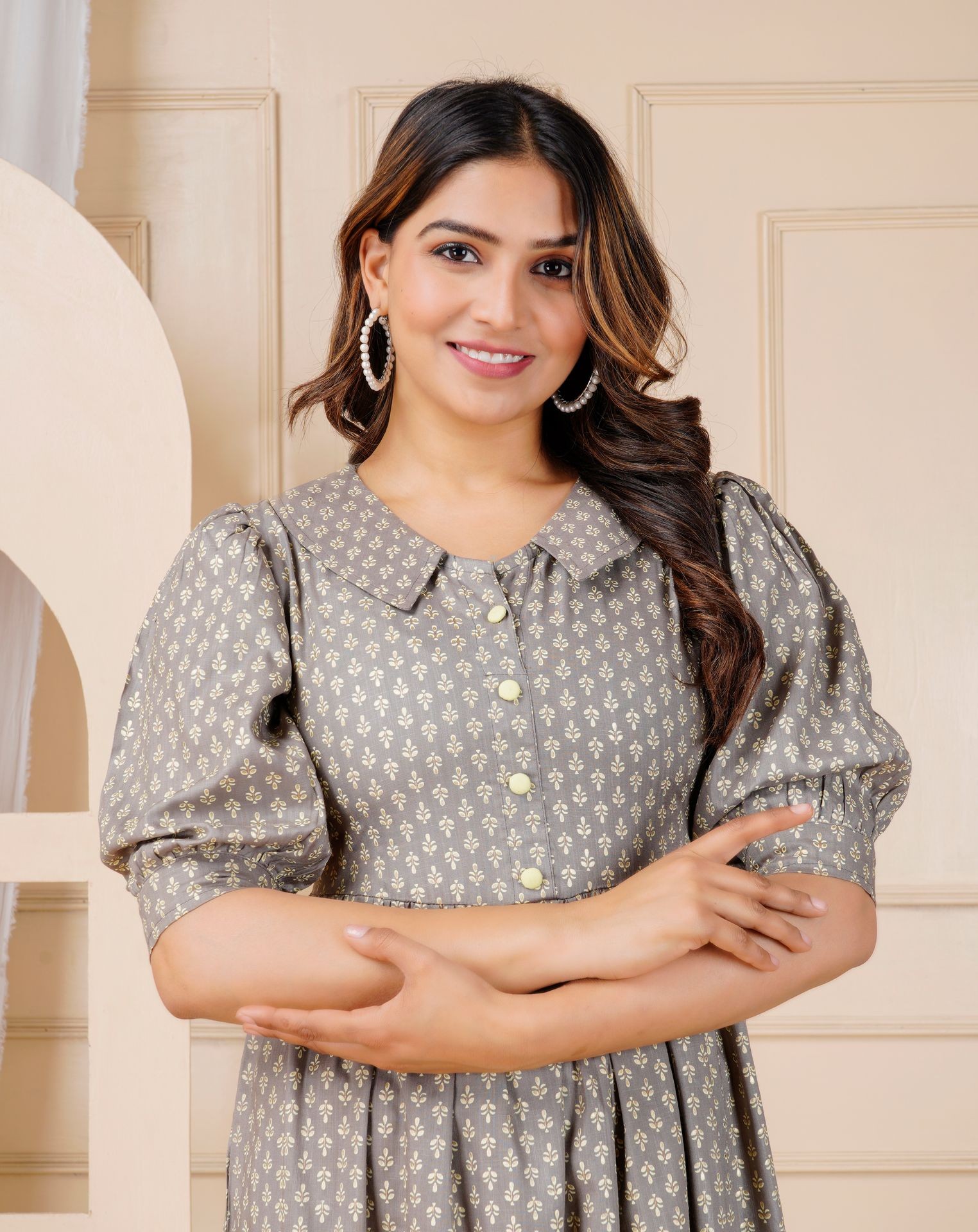 Woman in a gray dress with a floral pattern, standing against a beige wall, smiling.