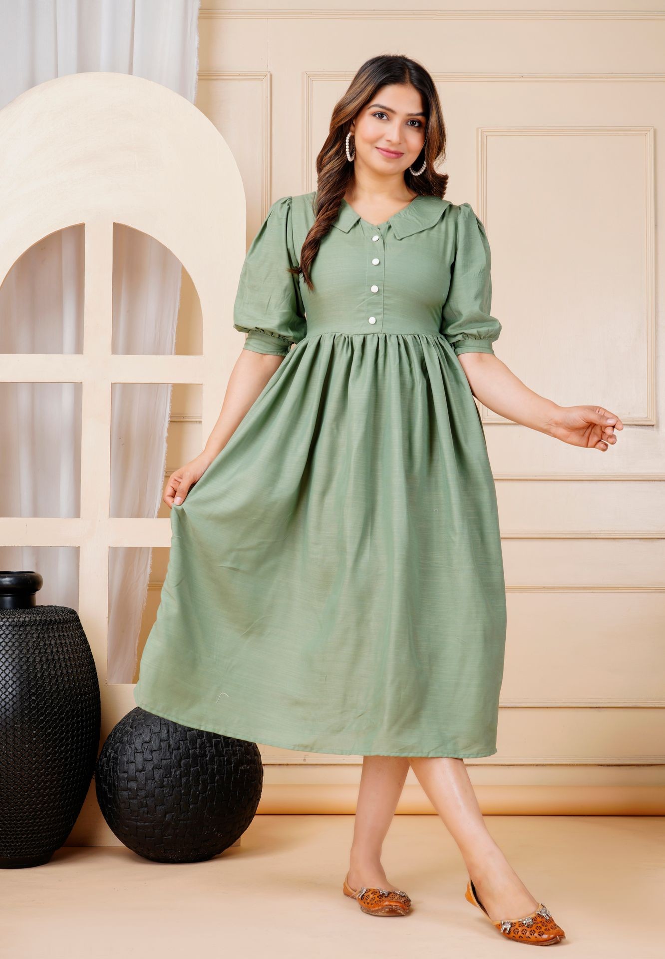 Woman in a green dress with puff sleeves and buttons, standing indoors by decorative vases.